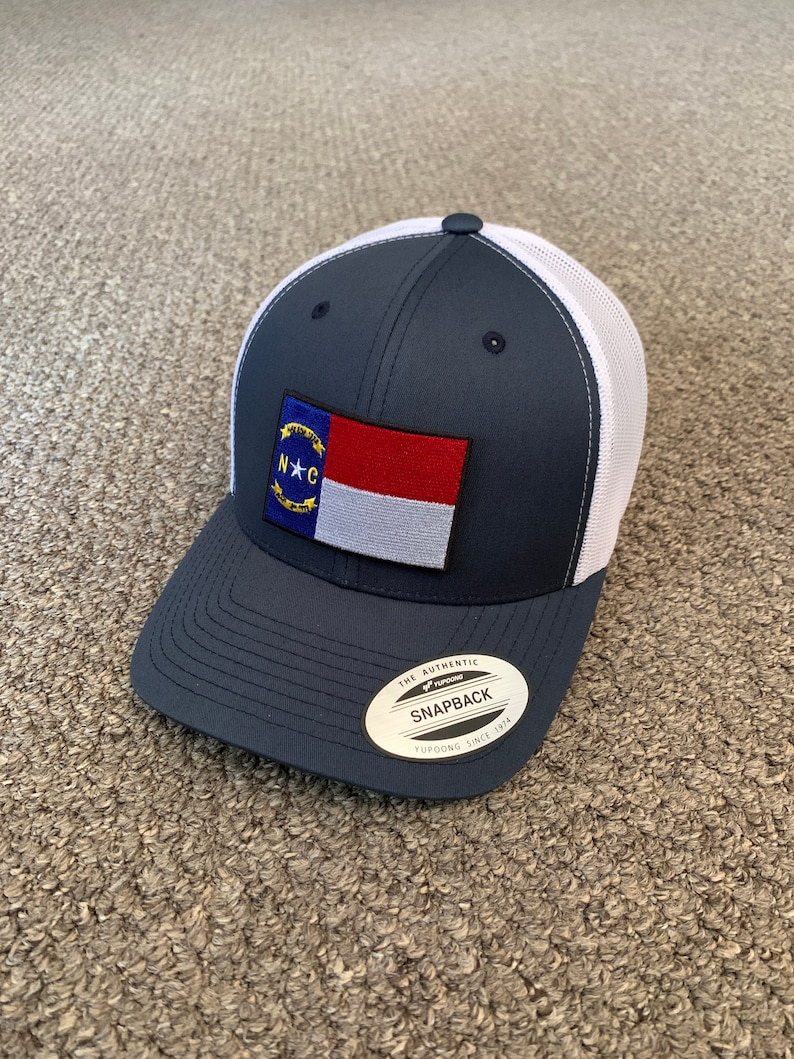 May include: A navy blue and white mesh trucker hat with a North Carolina state flag patch. The hat has a white mesh back and a navy blue front with a black "THE AUTHENTIC SNAPBACK YUPOONG SINCE 1974" sticker.