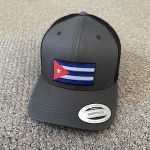 May include: A gray and black baseball cap with a Cuban flag patch on the front. The cap has a mesh back and a black snap closure. The label on the back reads "The Authentic Yupoong Snapback Yupoong Since 1974".