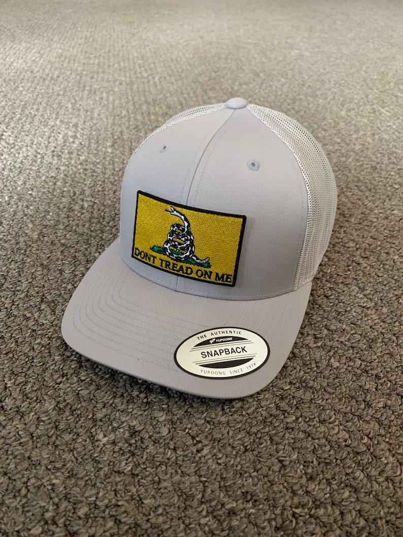 May include: Gray and white mesh trucker hat with a yellow patch featuring a coiled snake and the text "DON'T TREAD ON ME". The hat also has a silver label that reads "THE AUTHENTIC YUPOONG SNAPBACK SINCE 1974".