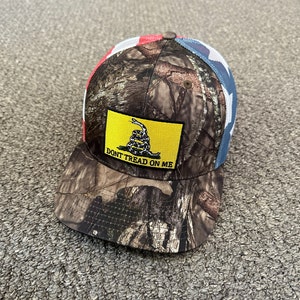 May include: A camouflage trucker hat with a red, white, and blue American flag mesh back. The front of the hat features a yellow patch with a coiled snake and the text "Don't Tread On Me".