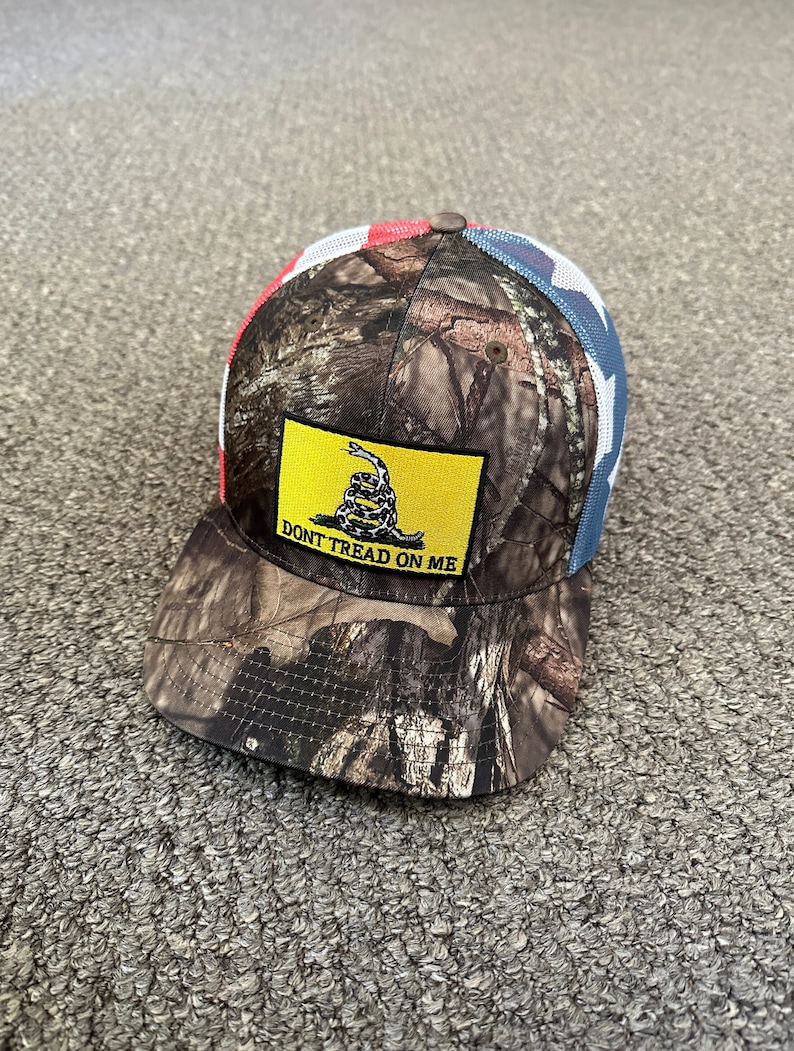 May include: A camouflage trucker hat with a red, white, and blue American flag mesh back. The front of the hat features a yellow patch with a coiled snake and the text "Don't Tread On Me".