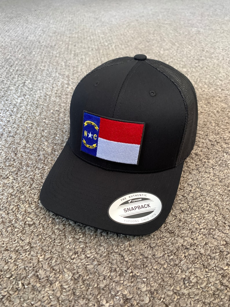 May include: A black and red North Carolina state flag patch on a black mesh trucker hat. The hat has a black brim and a black crown with a mesh back. The hat has a white label with the text "The Authentic Yupoong Snapback Yupoong Since 1974".