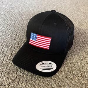 May include: Black and red American flag patch on a black mesh trucker hat with a black brim. The hat has a white label that says "The Authentic Yupoong Snapback Yupoong Since 1974".