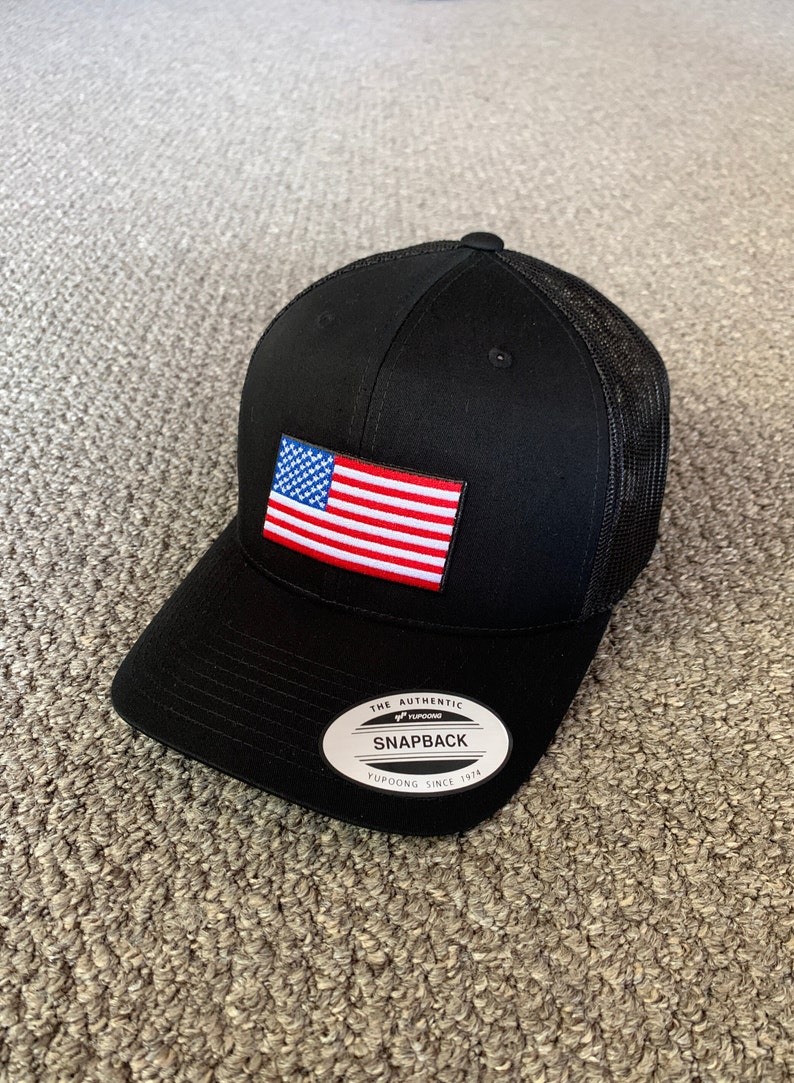 May include: Black and red American flag patch on a black mesh trucker hat with a black brim. The hat has a white label that says "The Authentic Yupoong Snapback Yupoong Since 1974".