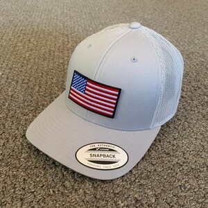 May include: A gray and white mesh trucker hat with an American flag patch and a black and white label that reads "The Authentic Yupoong Snapback Yupoong Since 1974".