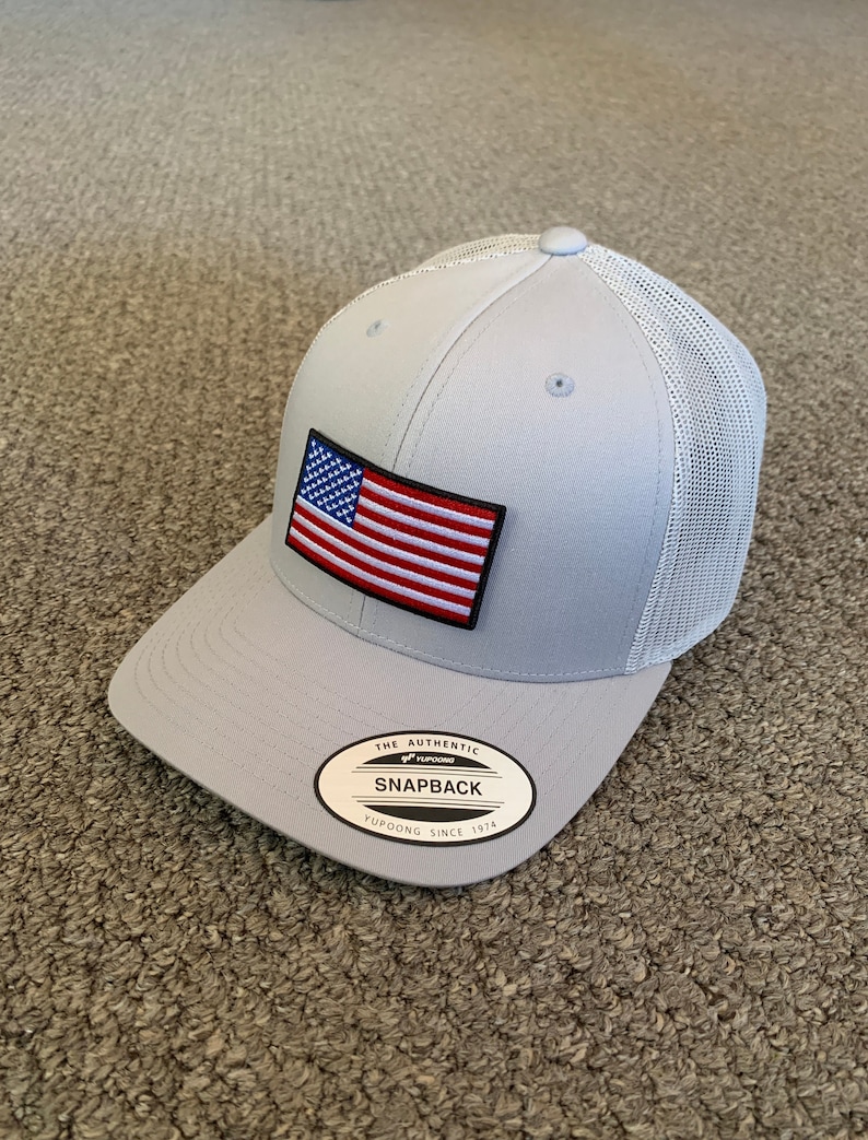 May include: A gray and white mesh trucker hat with an American flag patch and a black and white label that reads "The Authentic Yupoong Snapback Yupoong Since 1974".