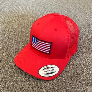 May include: Red and white American flag patch on a red mesh trucker hat with a black "The Authentic Snapback Yupoong Since 1974" label.
