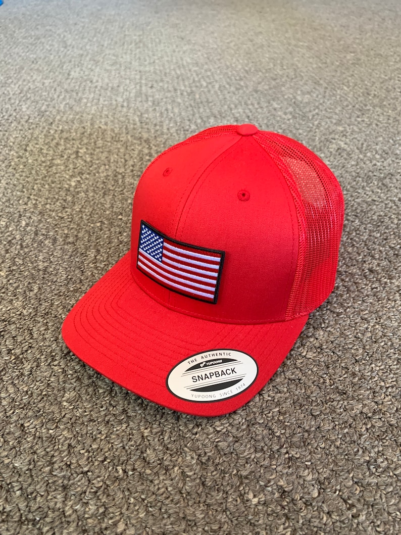 May include: Red and white American flag patch on a red mesh trucker hat with a black "The Authentic Snapback Yupoong Since 1974" label.