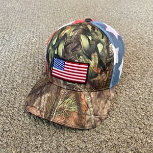 May include: A camouflage baseball cap with a red, white, and blue American flag patch on the front. The cap has a mesh back with white stars.
