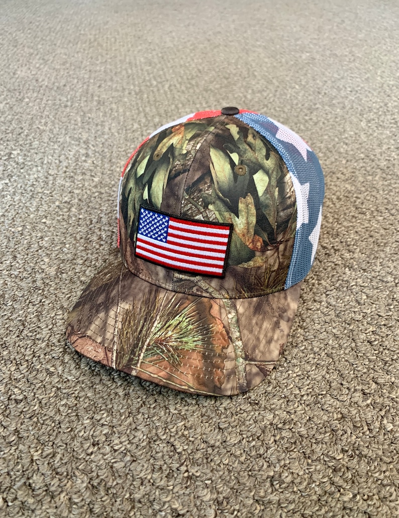 May include: A camouflage baseball cap with a red, white, and blue American flag patch on the front. The cap has a mesh back with white stars.