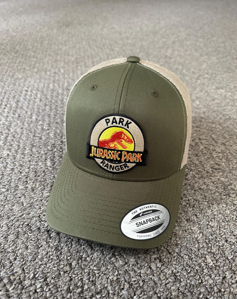 Puede incluir: Gorra de camionero verde y beige con un parche de logotipo de Jurassic Park Ranger. El parche presenta un c&iacute;rculo amarillo con un dinosaurio rojo y el texto "Jurassic Park Ranger". La gorra tambi&eacute;n tiene una etiqueta blanca que dice "The Authentic Snapback Yupoong Since 1974".