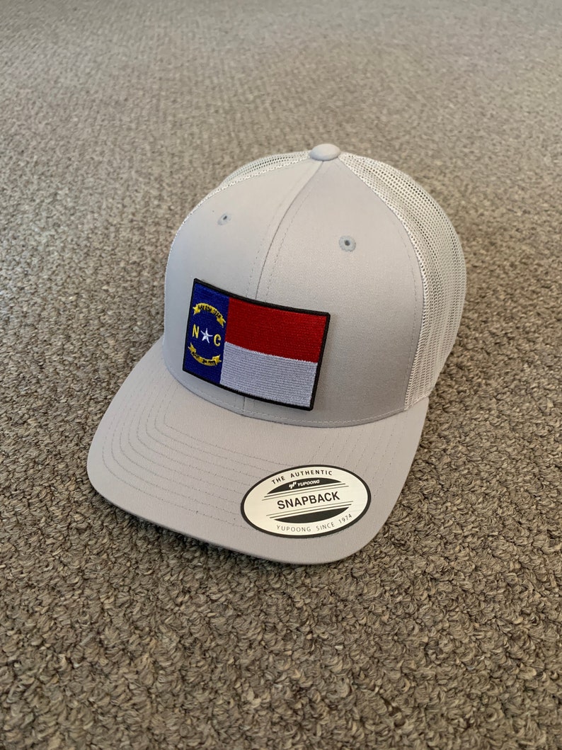 May include: Gray and white mesh trucker hat with a North Carolina state flag patch on the front. The hat has a black "The Authentic Yupoong Snapback Yupoong Since 1974" label on the back.