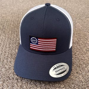 Może przedstawiać: Granatowa czapka typu snapback z białą siateczką z tyłu i usztywnionym panelem przednim. Haftowana naszywka z przodu przedstawia prostokątną flagę amerykańską z okręgiem gwiazd wokół liczby 250. Na daszku znajduje się srebrna naklejka.