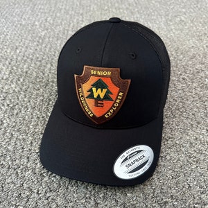 May include: Black mesh trucker hat with a brown patch that says "Senior Wilderness Explorer" with a green pine tree and a yellow "W" inside. The hat has a black brim and a white tag that says "The Authentic Yupoong Snapback Yupoong Since 1974."