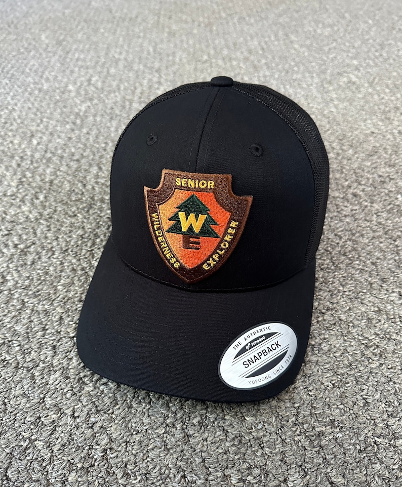 May include: Black mesh trucker hat with a brown patch that says "Senior Wilderness Explorer" with a green pine tree and a yellow "W" inside. The hat has a black brim and a white tag that says "The Authentic Yupoong Snapback Yupoong Since 1974."