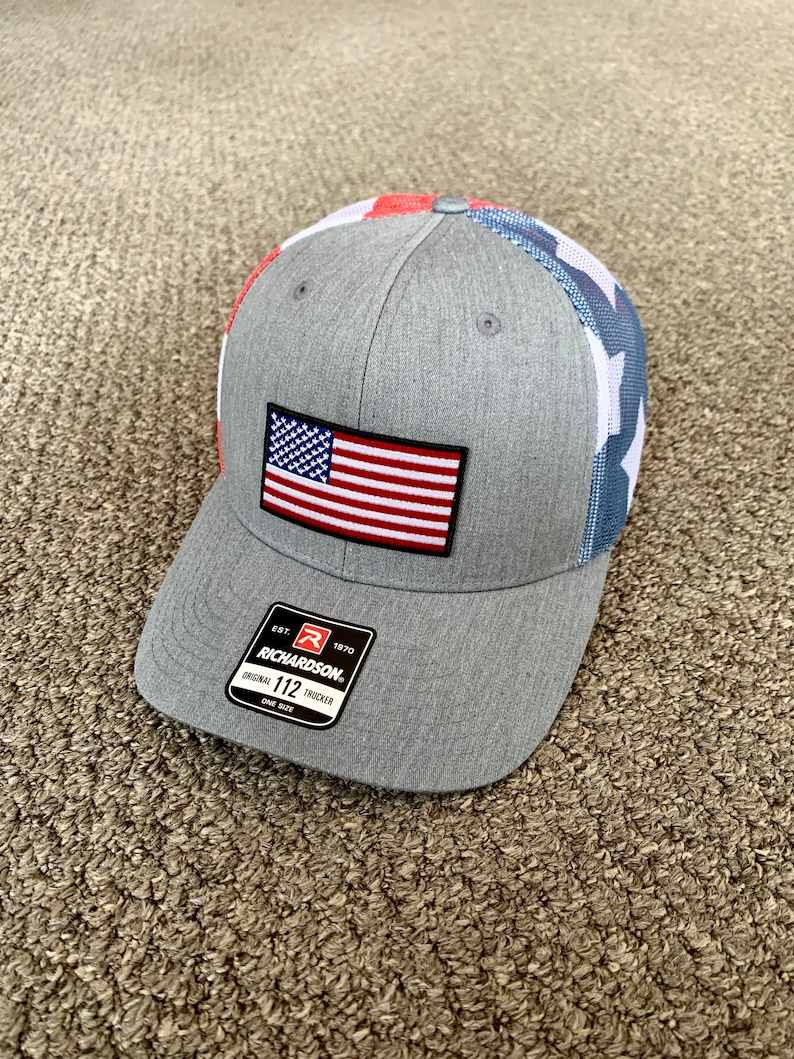May include: A gray and blue baseball cap with a red, white, and blue American flag patch on the front. The cap has a mesh back and a white "Richardson" logo with the text "Est. 1970" and "Original 112 Trucker" on the side.