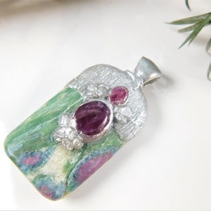 May include: A silver pendant with a green, pink, and white gemstone set in a silver frame. The gemstone has two pink ruby cabochons set in the silver frame.