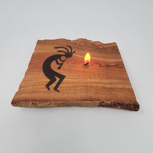 May include: A sandstone plaque with a black silhouette of a Kokopelli figure playing a flute. A small, lit candle sits on the plaque.