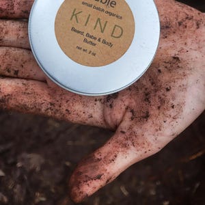 May include: A small, round, silver tin of "KIND" beard, babe, and body butter, net wt. 2 oz. The tin has a brown label with the product name and description. The tin is held in a hand covered in dirt.