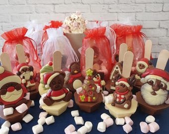 Op de afbeelding: Assortiment van kerstthema chocolade warme chocolademelk bommen met verschillende ontwerpen, waaronder Sinterklaas, een pinguïn, een teddybeer en een kerstboom. Elke bom is op een houten stokje en is omringd door witte marshmallows.