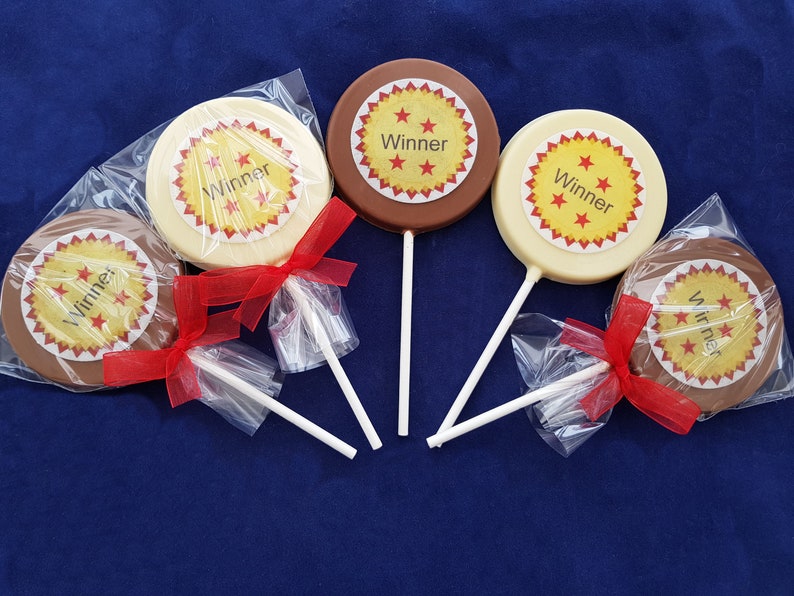 May include: Four round chocolate lollipops with a red, yellow, and white star design. The lollipops are wrapped in clear plastic and tied with a red ribbon. The lollipops say "Winner" on them.