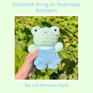May include: A crocheted green frog wearing blue overalls. The frog is holding a small flower in its hand. The frog is sitting on a green leafy background. The text "by LorettasLoops" is at the bottom of the image.