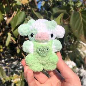 May include: A small green and white crocheted cow plush toy with a pink nose and black eyes. The cow is wearing a green crocheted overall with a heart-shaped patch.