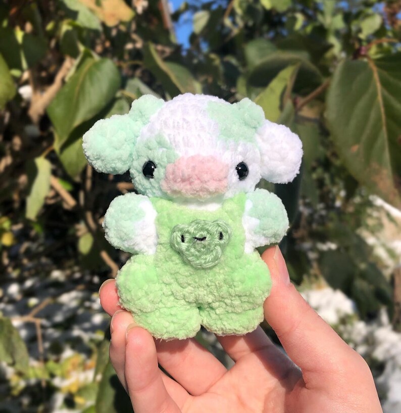May include: A small green and white crocheted cow plush toy with a pink nose and black eyes. The cow is wearing a green crocheted overall with a heart-shaped patch.