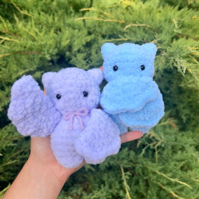 May include: Two small plush toys, one light blue and one lavender, are held in a hand. Both toys have black eyes and are made of a fuzzy material.