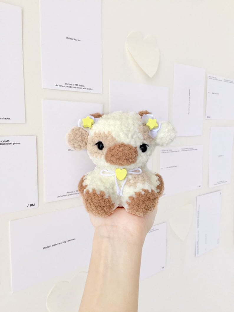 May include: A small crocheted plush toy cow with brown and white fur. The cow has yellow star-shaped ears and a yellow heart-shaped bow. The cow is sitting in a person's hand.
