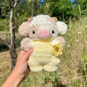 May include: A crocheted stuffed cow toy with a yellow and white spotted pattern. The cow is wearing a yellow crocheted outfit with a sunflower on the front.