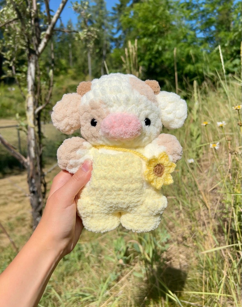 May include: A crocheted stuffed cow toy with a yellow and white spotted pattern. The cow is wearing a yellow crocheted outfit with a sunflower on the front.