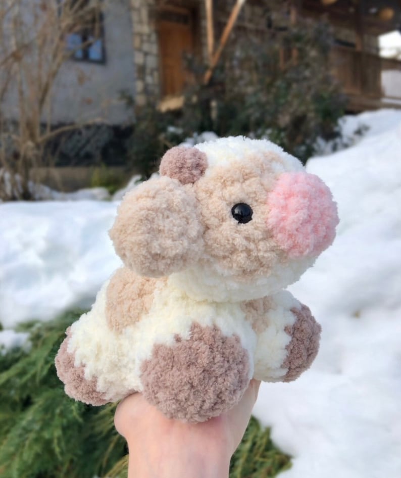 May include: A crocheted plush toy cow with brown and white patches. The cow has a pink nose and black eyes.