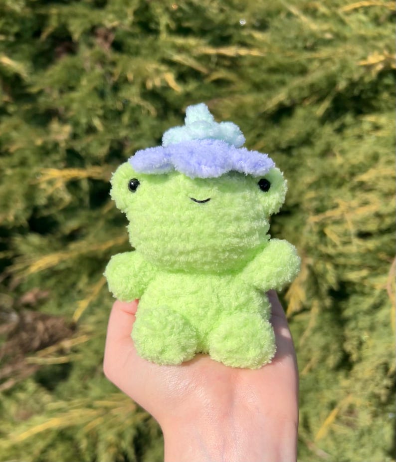 May include: A small green plush frog wearing a purple and light blue flower on its head.