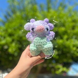 May include: A crocheted cow plushie with a purple and white body and green pants. The plushie has a pink nose and black eyes. The plushie is being held in a hand with a blurred background of green trees and a blue sky. The text "moo!" is written above the plushie and "@sunny.crochet" is written below the plushie.