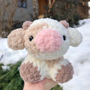 May include: A crocheted plush toy cow with brown and white patches. The cow has a pink nose and black eyes. The toy is sitting on a hand.