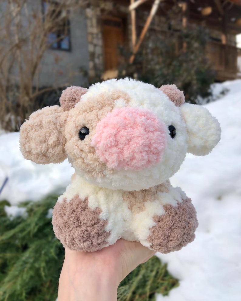 May include: A crocheted plush toy cow with brown and white patches. The cow has a pink nose and black eyes. The toy is sitting on a hand.