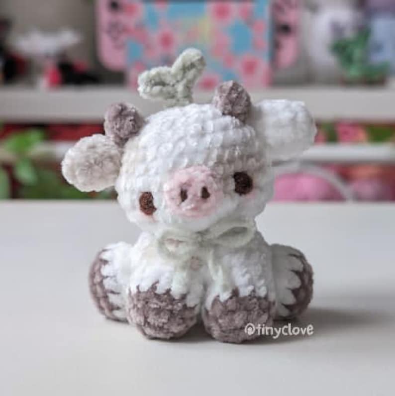 May include: A white crocheted cow plush toy with brown accents, a pink nose, and a green leaf on its head. The toy is sitting on a white surface.