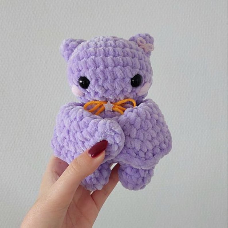 May include: A small, crocheted plush toy in the shape of a purple cat with a yellow bow. The cat has black eyes and a pink nose.