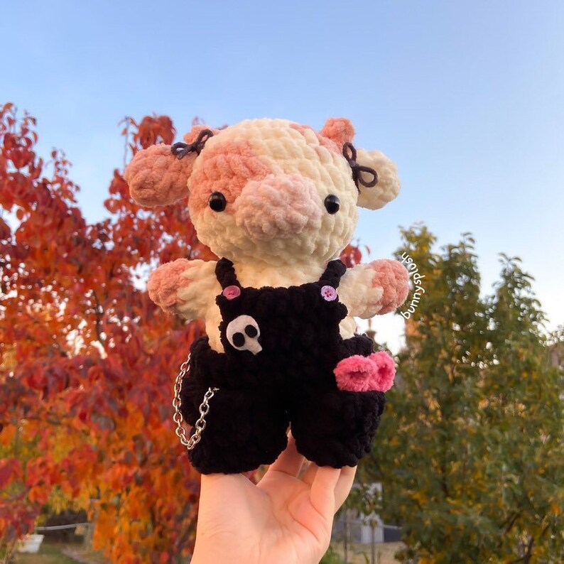 May include: A crocheted pink and white cow plush toy wearing black overalls with a skull and pink accents. The toy is being held in a hand against a background of fall foliage.
