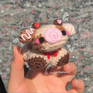 May include: A crocheted stuffed animal shaped like a pig with brown, white, and pink yarn. The pig has a pink nose, brown eyes, and a white swirl on its head. The pig is wearing a red ribbon around its neck.