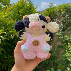 May include: A crocheted plush toy cow with black and white spots, wearing pink overalls with a brown button. The cow has a pink nose and black eyes.