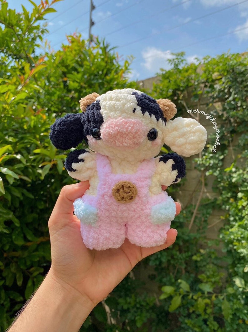 May include: A crocheted plush toy cow with black and white spots, wearing pink overalls with a brown button. The cow has a pink nose and black eyes.