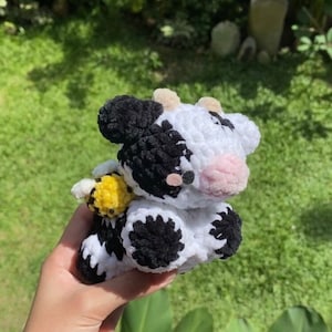 May include: A crocheted plush toy cow with black and white spots, holding a yellow and black crocheted bee. The cow has a pink nose and black eyes.