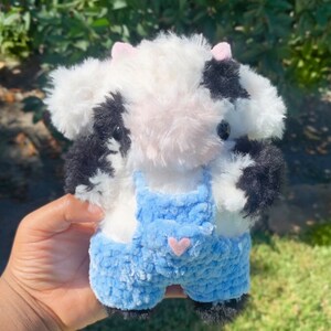May include: A small plush cow toy with black and white fur, wearing blue overalls with a pink heart on the pocket.