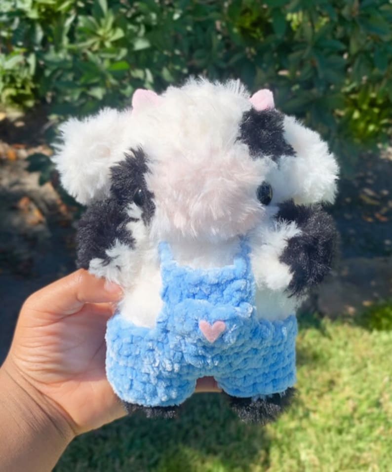 May include: A small plush cow toy with black and white fur, wearing blue overalls with a pink heart on the pocket.