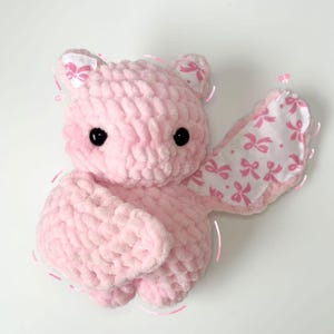May include: A pink crocheted bat plush toy with a white fabric wing featuring a pink bow pattern.
