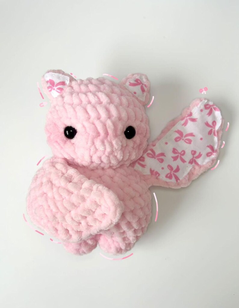 May include: A pink crocheted bat plush toy with a white fabric wing featuring a pink bow pattern.