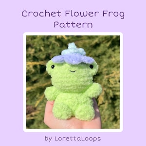 Może przedstawiać: Zielona dziana żaba z fioletowym i niebieskim kwiatem na głowie. Żaba siedzi w dłoni osoby. Wzór na dzianą żabę z kwiatem autorstwa LorettaLoops.