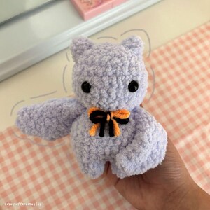 May include: A crocheted purple bat with orange and black ribbon. The bat has black eyes and a fluffy texture.